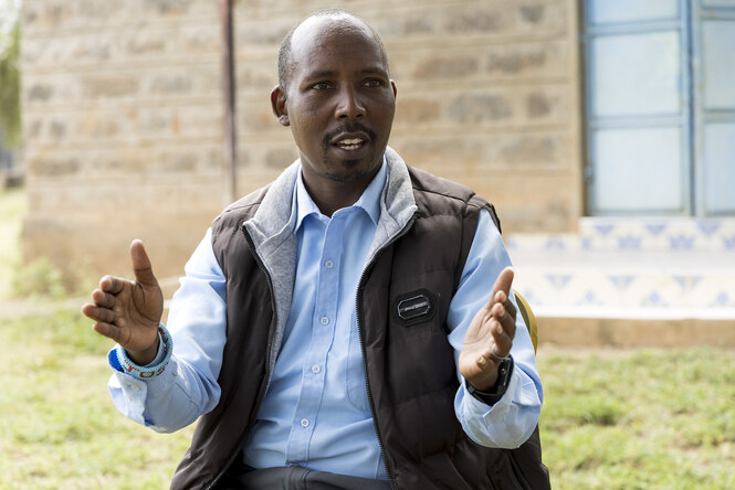 Porträt von Pastor Joseph Kiranto - er trägt ein hellblaues Hemd und eine braune Weste und erklärt seinem Gegenüber etwas