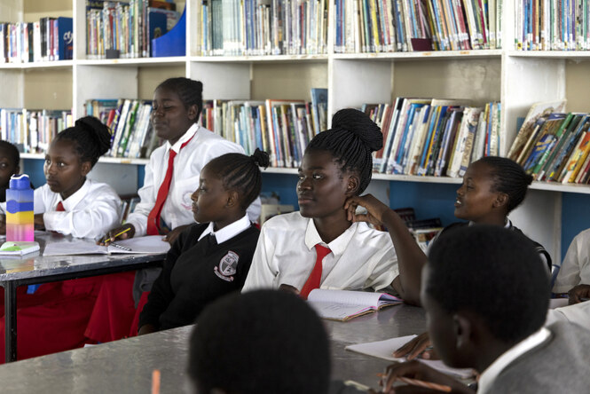 Schülerinnen in Schuluniform sitzen in einem Klassenzimmer und hören aufmerksam zu, im Hintergrund ein Bücherregal