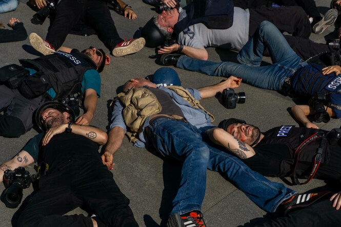 Leute mit Presse-Schutzwesten und Kameras liegen mit geschlossenen Augen auf dem Boden