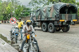 Motorradfahrer und ein Militaerfahrzeug auf einer Strasse in Cotonou, Benin