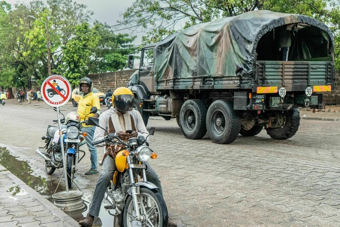Motorradfahrer und ein Militaerfahrzeug auf einer Strasse in Cotonou, Benin