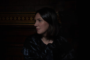 Claire M Singer vor einer Wand im Halbdunkel