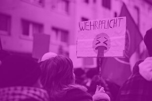 Menschen auf einer Demo; ein Schild mit dem Wort „Wehrpflicht“ über einem kotzenden Emoji wird hochgehalten.
