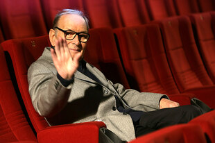 Ennio Moriccone winkt in die Kamera, er sitzt auf einem roten Plüschsessel im Kino