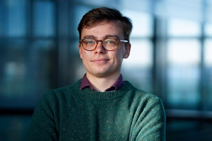 Porträt von Jonas Waack mit Hornbrille und petrolfarbenen Wollpullover. Unter dem Pullover trägt er ein lilanes Hemd, dessen Kragen zu sehen ist. Im Hintergrund ist verschwommen ein nicht näher bestimmbarer Raum mit Fenstern. Seine Mundwinkel könnten als schmunzelnd interpretiert werden.