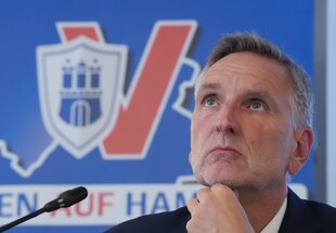 Torsten Voss sitzt an zwei Mikrofonen vor dem Logo des Hamburger Landesamts für Verfassungsschutz und guckt in die Luft