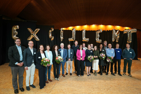 Menschen haben sich im Halbkreis aufgestellt, Sie halten Luftballon-Buchstaben, die, teils verdreht, das Wort "excellent" ergeben