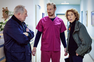 Ein Mann in lila Kittel steht zwischen den Ermittlern