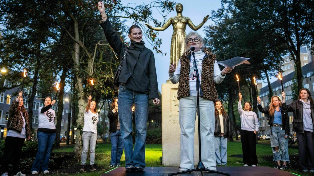Eine junge Frau und Dunya Verweij und stehen vor der Statue von Wilhelmina Drucker - die Frauen hinter ihnen im Halbkreis recken ihre brennenden Fackeln in die Höhe