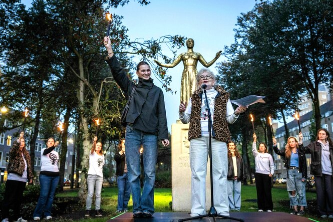 Eine junge Frau und Dunya Verweij und stehen vor der Statue von Wilhelmina Drucker - die Frauen hinter ihnen im Halbkreis recken ihre brennenden Fackeln in die Höhe