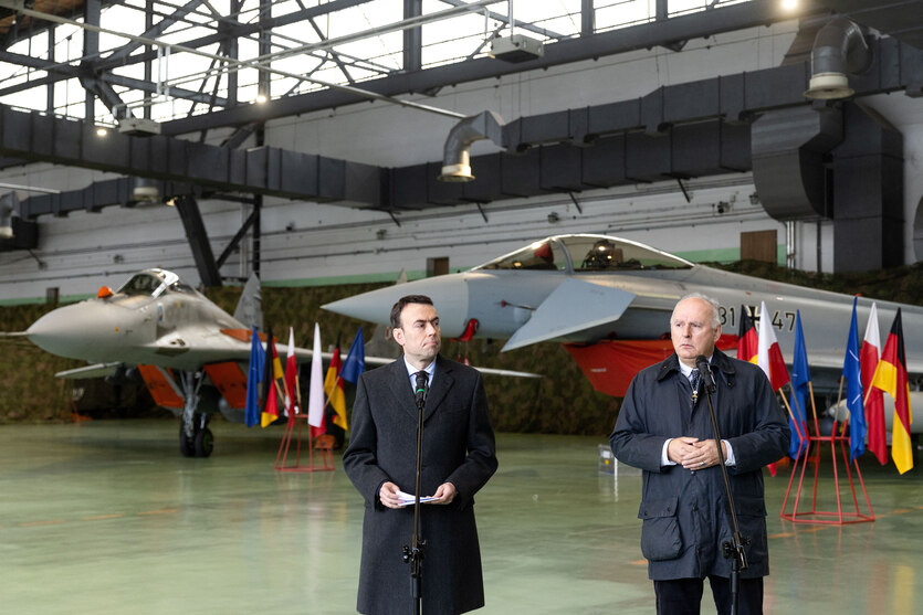Zwei Männer an Mikrofonen stehen in einer Halle, im Hintergrund parken Kampfjets