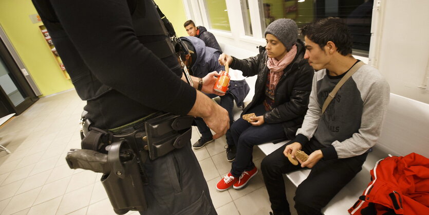 im Vordergrund des Bildes sieht man den Körper eines Polizisten, unterhalb des Kopfes, der vor zwei geflüchteten Jugendlichen steht, die auf einer Bank sitzen und bietet ihnen ein paar Kekse an. Im Hintergrund sieht man zwei andere junge Männer sitzen.