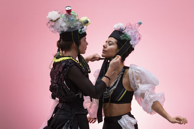 Vor rosa Hintergrund helfen sich zwei junge Frauen beim Anlegen einer "Tracht" aus gebrauchten Plastikgegenständen