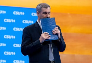 Markus Söder steht vor einer Wand mit einem CSU-Schriftzug-Muster. Er hält sich eine blaue Mappe vor dem Mund Markus Söder steht vor einer Wand mit einem CSU-Schriftzug-Muster. Er hält sich eine blaue Mappe vor dem Mund