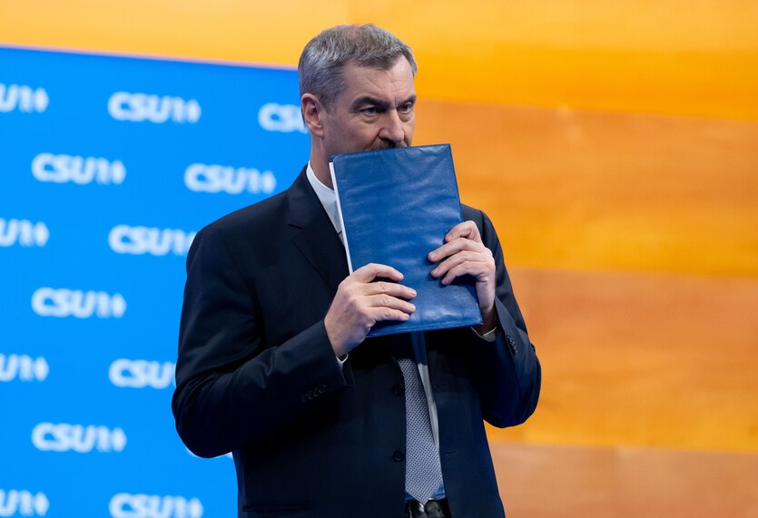 Markus Söder steht vor einer Wand mit einem CSU-Schriftzug-Muster. Er hält sich eine blaue Mappe vor dem Mund