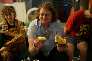 Drei Personen sitzen auf einem Sofa, die Person in der Mitte hält Gebäck in den Händen.