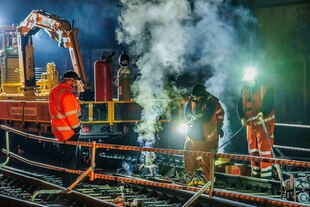 Ein Bautrupp ist im Bereich einer WeichenStörung im Einsatz. Es qualmt und dampft, die Männer tragen Warnkleidung und Bagger lauern im Hintergrund