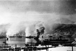 Auf einem alten Schwarzweißfoto sind brennende Schiffe in einem Hafen vor Bergen zu sehen