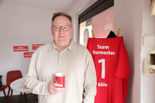 Burmester schaut mit einer Tasse in der Hand in die Kamera. Im Hintergrund ein Shirt mit der Nr.1 und der Aufschrift Team Burmester