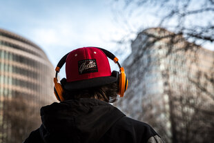 Ein Jugendlicher mit orangen Kopfhörern und Basecap von hinten, im Hintergrund Hochhäuser