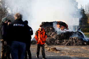 Ein Bauer in roter Jacke steht vor einem brennenden Haufen mit Strohballen