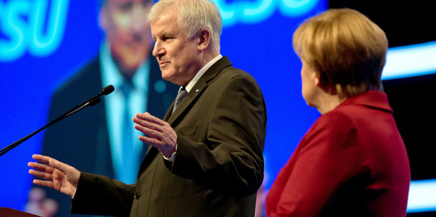 Horst Seehofer steht mit ausgebreiteten Armen an einem Rednerpult, neben ihm steht Angela Merkel und hört ihm zu.
