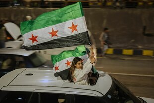 Ein Mädchen schaut aus einem Autodach heraus, sie hält eine Fahne Syriens in den Händen, die im Fahrtwind weht