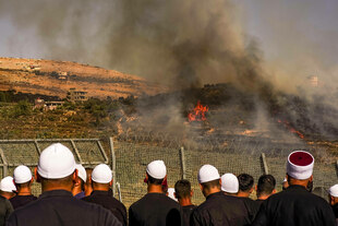 Männer mit weißen Kopfbedeckungen stehen vor einem Zaun, auf dem Gelände dahinter brennt es, die Männer blicken auf den Brand
