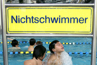 Ein Schild mit der Aufschrift „Nichtschwimmer“ hängt über dem Beckenrand eines Hallenbads. Dahinter plantschen mehrere Kinder im flachen Wasser, teilweise mit Schwimmhilfen Ein Schild mit der Aufschrift „Nichtschwimmer“ hängt über dem Beckenrand eines Hallenbads. Dahinter plantschen mehrere Kinder im flachen Wasser, teilweise mit Schwimmhilfen