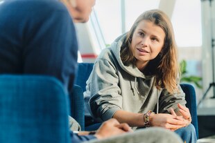 Zu sehen ist die Aktivistin und Autorin Luisa Neubauer in einem grünen Hoodie. Sie sitzt auf einem blauen Sessel, links vor ihr ist eine Schulter und ein Kinn einer Person zu erkennen.