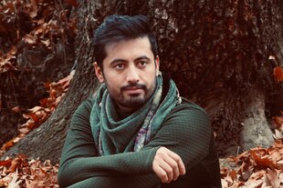 Porträt von Jafar Najafi mit Bart und kurzen Haaren - er sitzt auf dem mit Laub bedeckten Bodenan einem Baumstamm. Das Halstuch passt zu seinem grünen Pullover.
