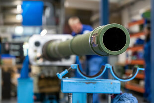 Nahaufnahme einer Kanone an der gearbeitet wird - die Arbeiter im Hintergrund der Werkstatt sind nur unscharf zu erkennen