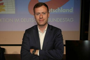 Porträt von Jan Wenzel Schmidt – er steht im Anzug mit verschränkten Armen vor einer Wand mit einer Projektion des AfD-Logos