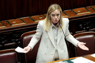 Giorgia Meloni steht im Parlament in Rom - Holz und Lederambiente - sie macht eine ratlose Geste während sie spricht. Meloni trägt einen hellen Hosenanzug.