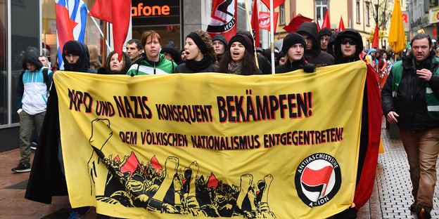 Im Vordergrund ein Banner, auf dem steht: "NPD und Nazis konsequent bekämpfen!" und weiter "Dem völkischen Nationalismus entgegentreten!". Das Banner tragen AntifaschistInnen, dahinter läuft ein bunter Demonstrationszug durch die Stadt.