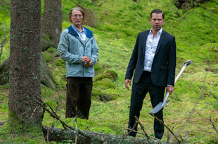 Mads Mikkelsen und Nikolaj Lie Kaas stehen in einem Wald mit leuchtend grünem Gras Mads Mikkelsen und Nikolaj Lie Kaas stehen in einem Wald mit leuchtend grünem Gras