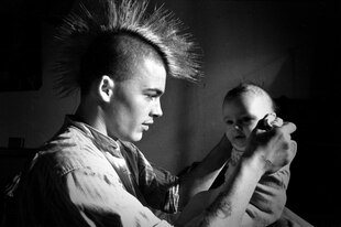 Ein schwarz-weißer Filmstill, auf dem ein junger Mann mit Irokesenschnitt ein Baby hält