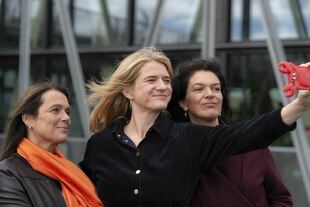 Drei Frauen stehen im Freien vor einer modernen Glasfassade und machen gemeinsam ein Selfie mit einem Smartphone in einer auffälligen, roten Krebs-Hülle. Von links nach rechts: Barbara Junge mit dunkler Lederjacke und leuchtend orangefarbenem Schal, Katri