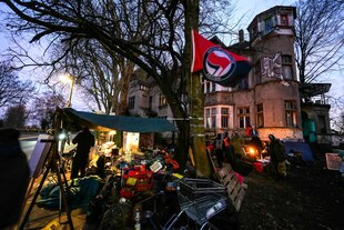 Vor der Villa Kunterbunt - ein altes Haus mit Erkern weht die Flagge der Antifa, davor stehen Menschen, ein Zelt auf dem Bürgerssteig und Habseligkeiten in Einkaufswagen