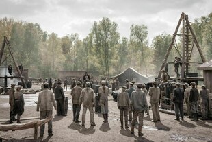 Szene aus einer historischen TV-Serie mit vielen Menschen, die zwischen Ölförderanlagen stehen.