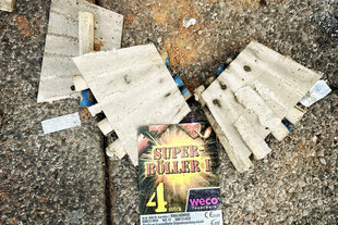 Pappreste von Feuerwerksraketen liegen auf einer Straße, aufgenommen von oben. Auf dem noch lesbaren Teil einer Pappverpackung steht "Superknaller 4" und der Name der Firma Weco. Neben der Pappe auf dem Boden ist rötlicher Staub vom Abfeuern der Raketen.