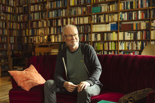 Ein Mann sitzt in einem Zimmer vor einem Regal mit sehr vielen Büchern