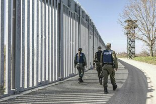 Ein hoher Grenzzaun und Uniformierte Bewacher:innen