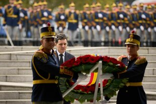 Zwei Soldaten in Parade-Uniform tragen einen Blumenkranz, dahinter läuft ein Mann im Anzug. Im Hintergrund stehen weitere Soldaten in Parade-Uniform auf einer steinernen Treppe.