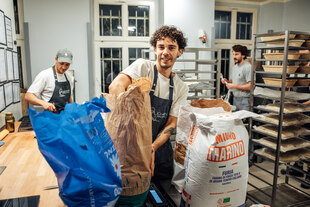 Szene in der Backstube der Bäckerei Johann -drei junge Männer in Arbeitskleidung - einer von ihnen greift in einen Mehlsack