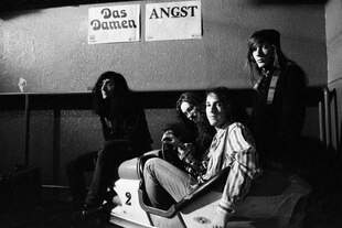 Vier junge Männer mit langen Haaren von der Band Das Damen posieren. Hinter ihnen hängen zwei Plakate, die Konzerte von Das Damen und Angst ankündigen