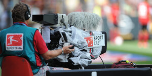 Ein Kameramann sitzt während eines Fußballspiels hinter seinem monströsen Aufnahmegerät und beobachtet das Spielgeschehen durch die Linse seiner Kamera.