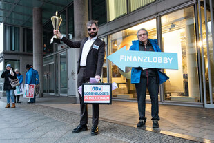 Eine Person steht im Anzug vor einem Gebäude, er hält einen Koffer in der Hand und einen Pokal in der anderen. Neben ihm hält eine weitere Person ein Plakat mit dert Aufschrift "Finanzlobby". Das Plakat hat die Form eines Pfeils, der auf den Mann im Anzug