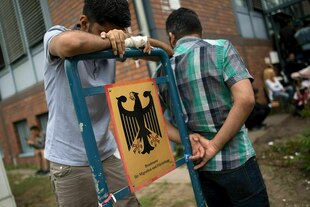 Zwei Männer stehen vor einem Amtsgebäude, einer lehnt an einem Schild mit einem Bundesadler. Zwei Männer stehen vor einem Amtsgebäude, einer lehnt an einem Schild mit einem Bundesadler.