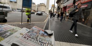 Zeitungen im Vordergrund, im Hintergrund laufen Menschen eine Straße entlang
